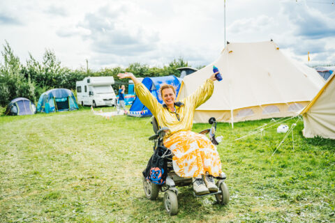 A smiling person in a yellow outfit sits in a motorised wheelchair, raising their arms joyfully at an outdoor campsite with tents, grass, and cloudy skies in the background.