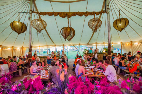 People sit at round tables eating and talking under a large, decorated marquee with hanging rustic lanterns. The scene is colourful and festive, with plants and flowers in the foreground.