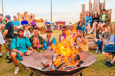 A group of people in colourful, festival-style clothing sit and stand around a large fire pit outdoors, with others in the background enjoying a lively, sunny festival atmosphere.