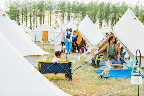 Children play among tents at a grassy campsite at Shambala. One child pulls another in a wagon, whilst adults and other children gather near neighbouring pre-pitched camping tents, with trees in the background.
