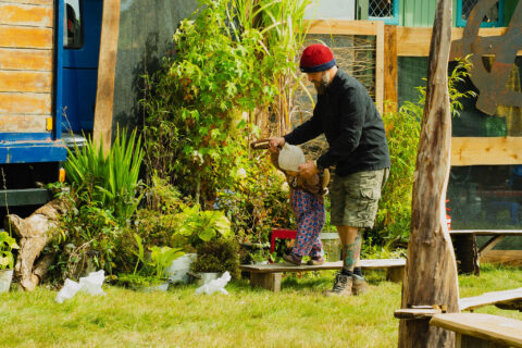 A man in a red beanie helps a small child wearing patterned trousers on a wooden bench in a green garden area, surrounded by plants and wooden structures.