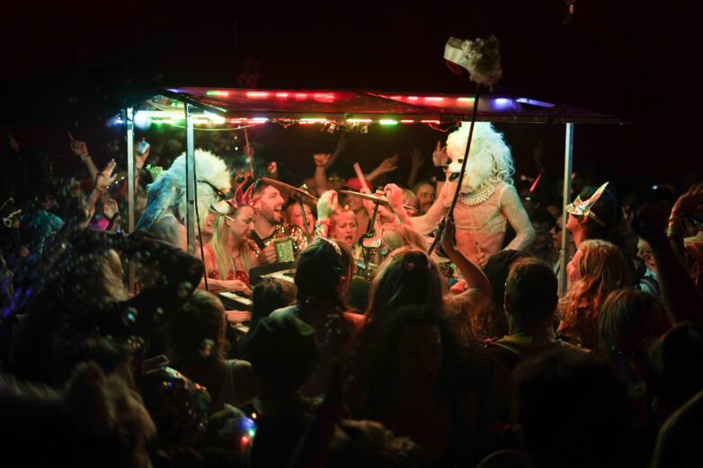 A crowded party scene with people in fancy dress and colourful outfits dancing around a stage with performers wearing furry white masks, under vibrant, multicoloured lights.