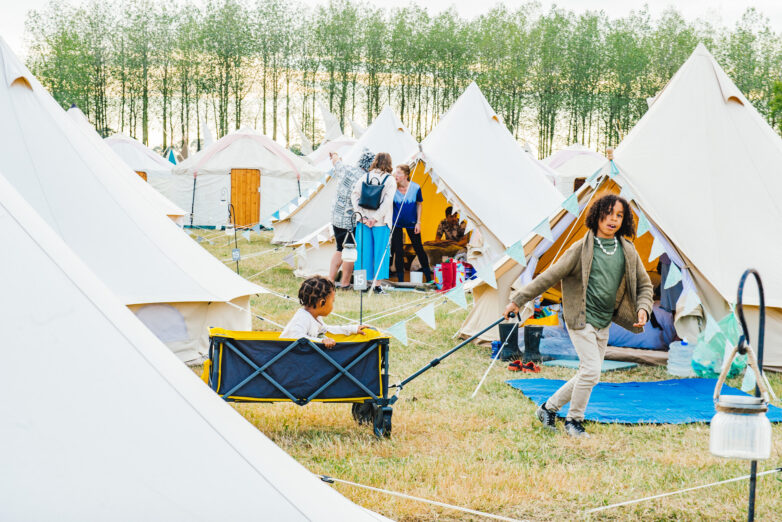 Children play among rows of cream-coloured tents at a campsite. One child sits in a yellow wagon while another pulls it. Adults and children gather near tents in the background. Trees line the horizon behind the camp.
