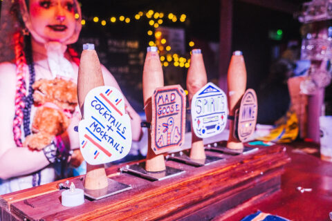 A row of wooden beer taps labelled with hand-drawn signs for Cocktails + Mocktails, Mad Goose, Spirits, and Wine, with a person wearing a fancy dress costume in the blurred background.