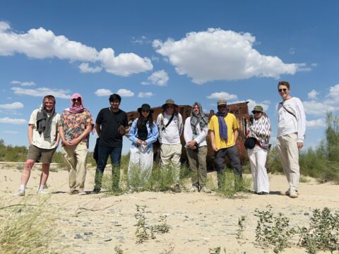 Nine people stand in a sunny, sandy area with scattered plants, dressed in casual and outdoor clothing. A rusted metal structure is in the background. The sky is blue with fluffy clouds.