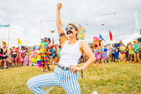 A person in a fake moustache, white vest, and blue striped trousers strikes a pose in front of a cheering crowd at Shambala festival. People wear colourful costumes and wigs. The sky is cloudy.