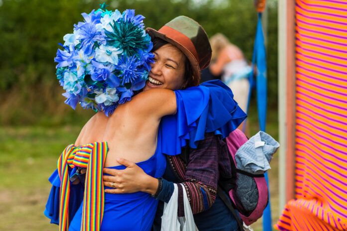 Two people hug and smile outdoors; one wears a large headpiece made of blue flowers and a bright blue dress with a rainbow-striped ribbon, while the other wears a brown hat and carries a bag.