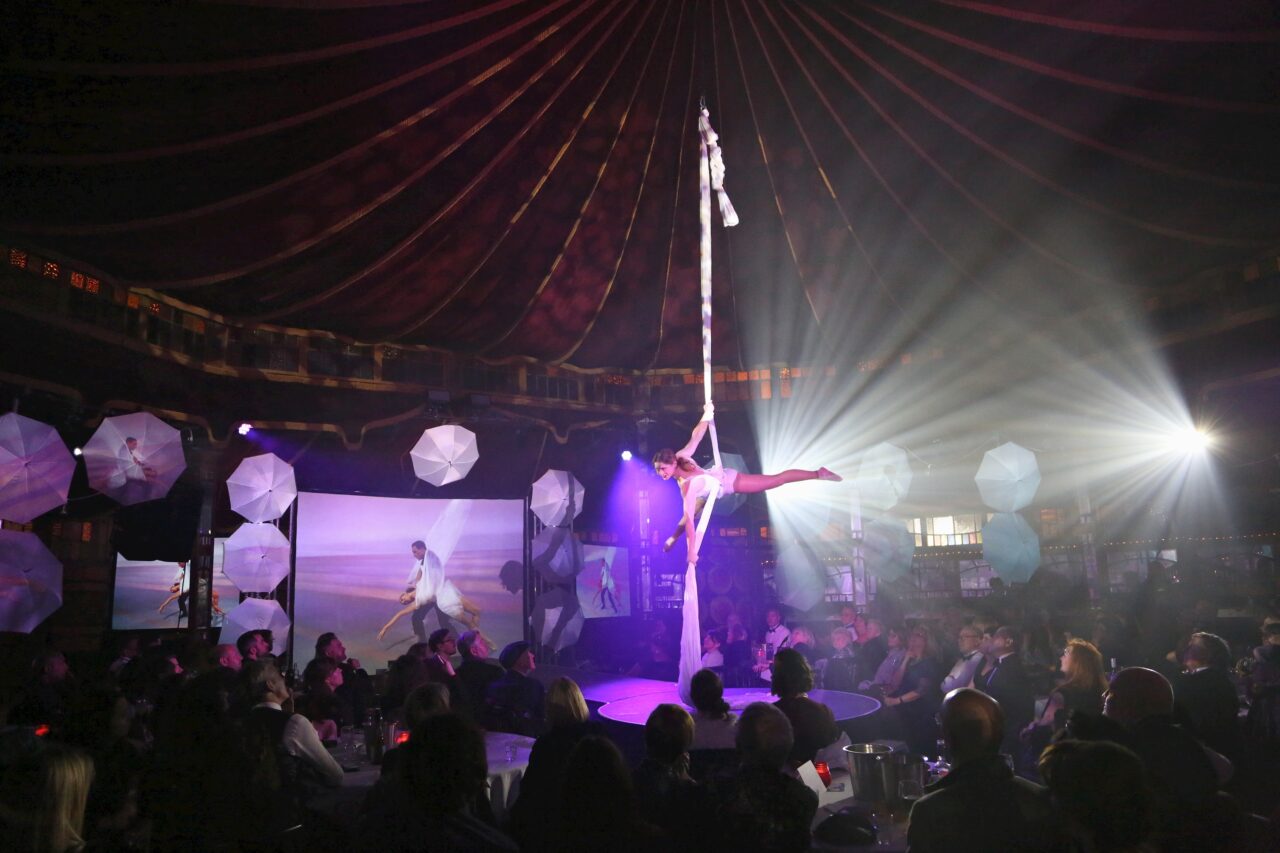 Aerial performer in a white costume hangs from fabric, suspended above a crowd seated at round tables under a Spiegeltent with dramatic stage lighting and large decorative parasols in the background.