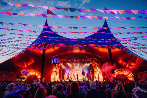 A large crowd watches a vibrant music performance under a marquee decorated with colourful bunting, illuminated by dramatic stage lighting at dusk.
