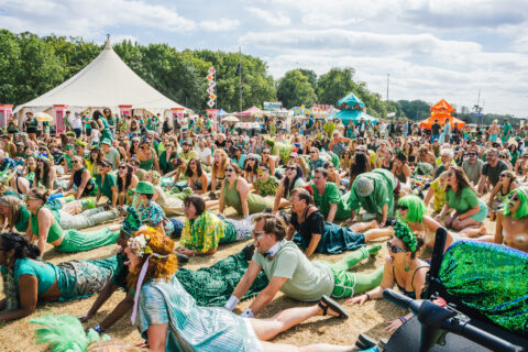 A large crowd of people dressed in green outfits participate in a group activity outdoors at Shambala, with tents and colourful decorations in the background under a partly cloudy sky.