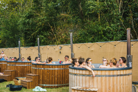 Several groups of people relax in large wooden hot tubs outdoors, enjoying drinks and conversation. Trees and a tan privacy fence are in the background, with towels and baskets nearby on the grass.
