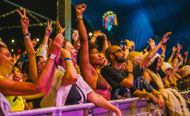 A diverse crowd of people at a main stage performance, standing behind a barrier, raising their arms, cheering, and enjoying the vibrant atmosphere under colourful lights.