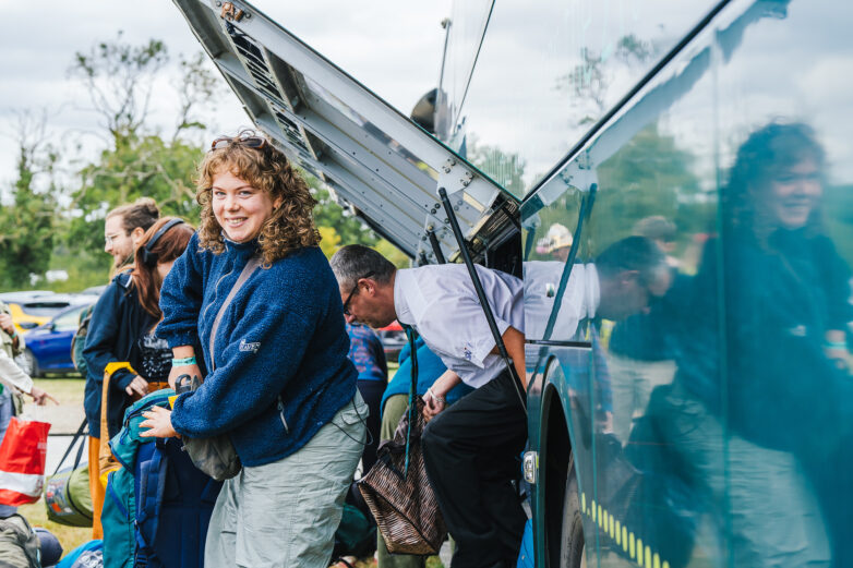 A smiling woman stands near a coach with an open luggage compartment, holding a blue sleeping bag. Other people are unloading bags and items from the coach in the background. Trees and cars are visible in the distance.