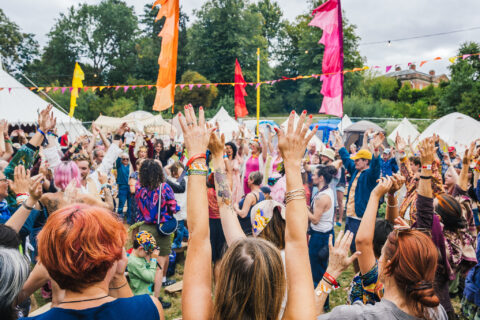 A large group of people at Shmabala festival raise their hands in the air, surrounded by colourful flags, bunting, and tents, with trees and a cloudy sky in the background.