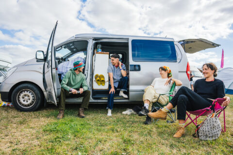 Four people relax by a silver campervan on a grassy field, talking and smiling. Two sit inside the van, two on camping chairs outside. The van doors are open, and the sky is partly cloudy.