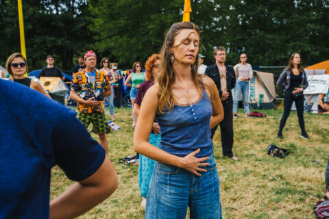 A woman stands outdoors with her eyes closed and hand on her stomach, appearing to meditate or focus on her breathing. Several other people stand around her on the grass, some also with eyes closed. Trees and tents are in the background.