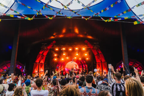 A large crowd with raised hands enjoys a live music performance on a brightly lit stage, decorated with colourful lights and flags, under a tented roof at Shambala festival.