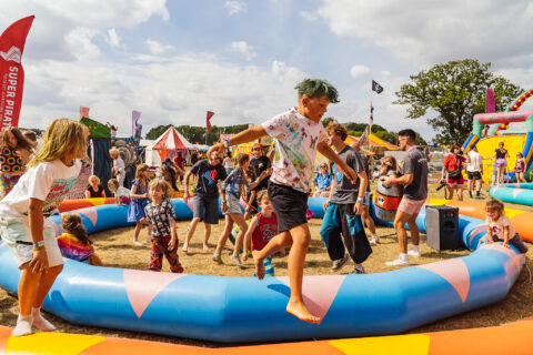 Children and adults play and jump inside an inflatable arena at Shambala, surrounded by colourful tents and flags on a sunny day.