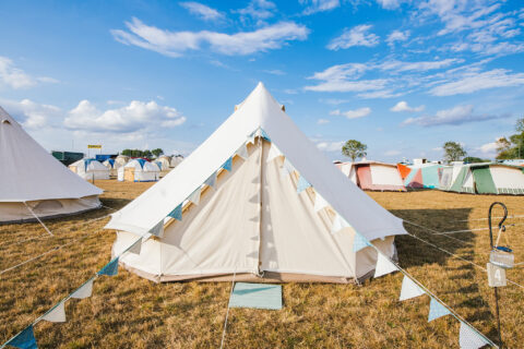 A white canvas tent decorated with blue and white pennant bunting stands on a grassy field among other tents under a bright blue sky with scattered clouds.