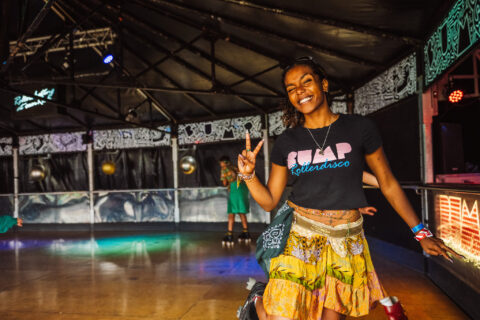 A smiling woman on roller skates flashes a peace sign in a vibrant, indoor roller rink. She wears a BUMP Rollerdisko T-shirt, yellow patterned skirt, and colourful accessories. The background features disco balls and bright lights.