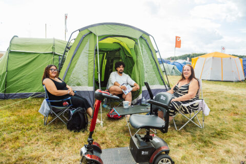 Three people sit on folding chairs outside a large green tent at a campsite, smiling at the camera. A mobility scooter is parked in front of them, and other tents are visible in the background.