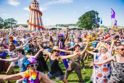 A large crowd of people in colourful, festive costumes dance outdoors at Shambala. A striped funfair helter-skelter and marquees are visible in the background under a bright blue sky.