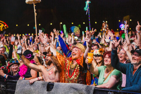 A lively crowd at the front row of Shambala Festival, many dressed in colourful costumes and accessories, cheer and raise their hands.