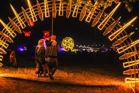 Two people walk through a glowing, circular arch of yellow lights at night during a festival, with illuminated decorations and marquees visible in the background.
