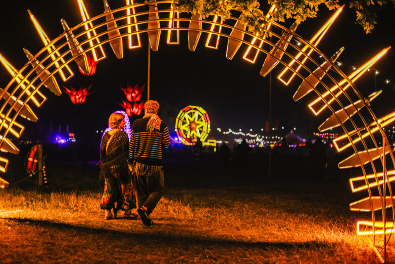 Two people walk through a glowing, circular arch of yellow lights at night during a festival, with illuminated decorations and marquees visible in the background.