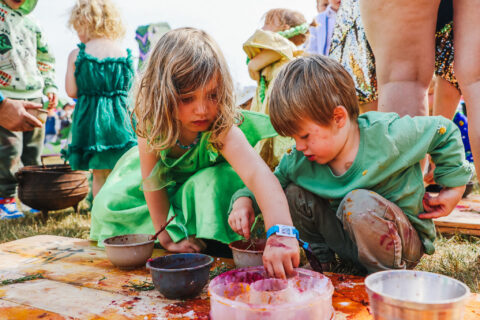 Two young children sit on the grass, wearing green clothes, reaching into bowls of colourful paint or food, participating in an outdoor activity surrounded by other children and adults.