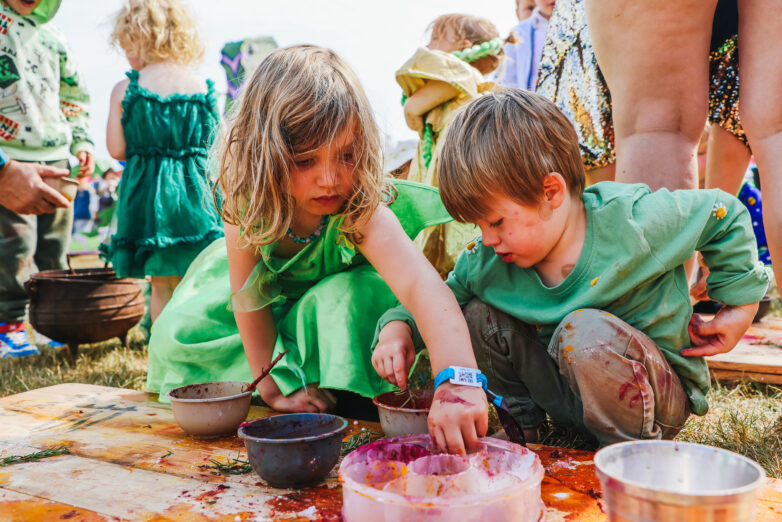 Two young children sit on the grass, wearing green clothes, reaching into bowls of colourful paint or food, participating in an outdoor activity surrounded by other children and adults.