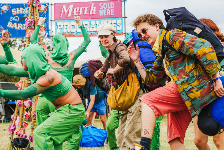 People in colourful costumes and outfits dance through the Shambala gates. Two signs in the background read Merch Sold Here and Programmes.