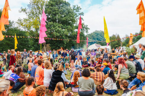 A large group of people sit in a circle outdoors at Shambala festival, surrounded by vibrant flags and trees, while others stand and watch. Tents and decorations are visible in the background under a partly cloudy sky.