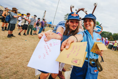 Two people in quirky costumes hand out colourful envelopes at the Shambala Carnival. Both are smiling; one wears a blue hat and glasses, the other has a hat with large googly eyes. Festival-goers mingle in the background.