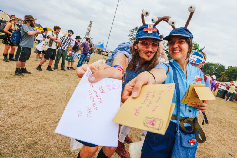 Two people in quirky costumes hand out colourful envelopes at the Shambala Carnival. Both are smiling; one wears a blue hat and glasses, the other has a hat with large googly eyes. Festival-goers mingle in the background.