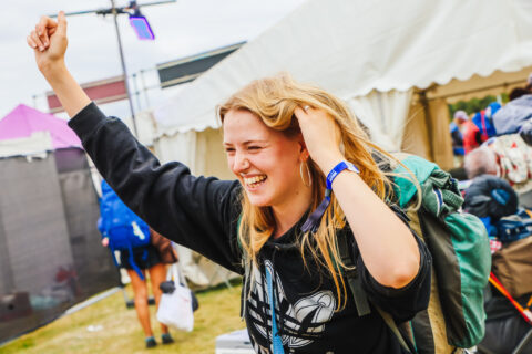 A young woman with a rucksack smiles joyfully and raises her fist in the air at an outdoor event, standing in front of white marquees, surrounded by people arriving from shuttle buses and trains.