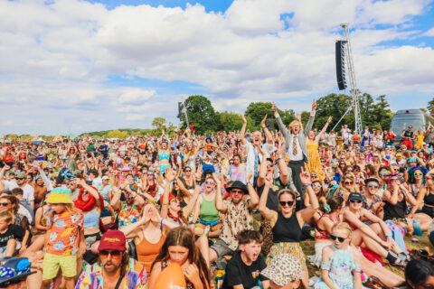 A large, colourful crowd of people gathered outdoors at Shambala during the day, many smiling, cheering, and raising their hands, with trees, blue sky, and clouds in the background.