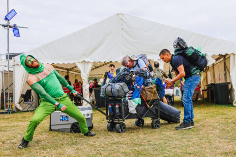 A person in a bright green costume strikes a dramatic pose next to people pulling luggage and camping equipment outside a large white marquee at at the entrance to Shambala