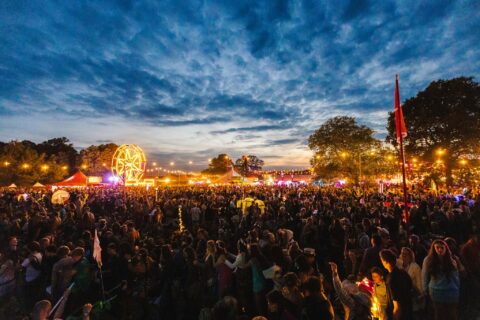 A large crowd gathers at Shambala at dusk, with fairy lights, trees, a Ferris wheel and colourful tents illuminating the lively scene under a dramatic, partly cloudy sky.