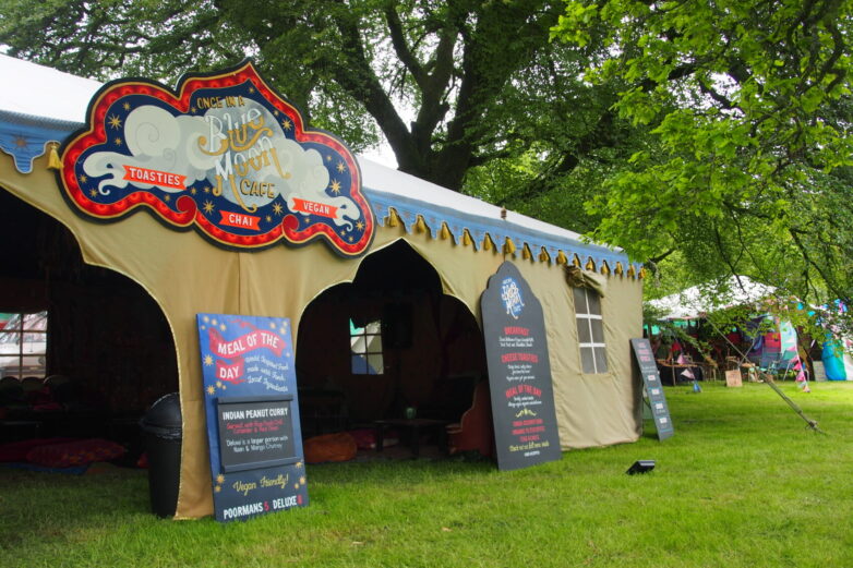 A large yellow tent, labelled Blue Moon Café, stands on grass under trees. Menu boards and a colourful sign advertise toasties, chaat, and vegan meals. Cushions are visible inside the tent.