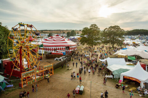 A lively outdoor festival scene with a Ferris wheel, a large red and white striped circus tent, and various stalls captures the vibrant spirit inspired by Shambala Festival history under a partly cloudy sky at sunset.