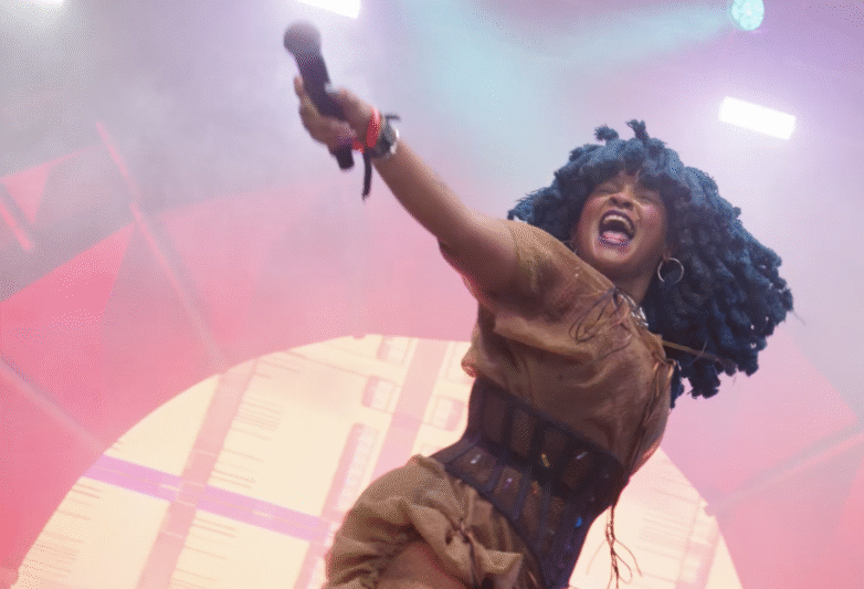 A performer with blue curly hair sings passionately on stage, holding a microphone and extending one arm. Bright stage lights and a colourful, smoky background create an energetic concert atmosphere.