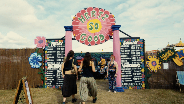 Three people walk through a colourful festival entrance with a flower-shaped sign reading Hertz So Good and timetable boards for Friday, Saturday, and Sunday on either side. Other attendees and tents are visible in the background.