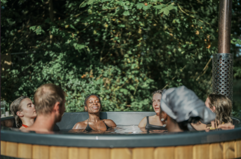 Six people relax together in a round outdoor hot tub surrounded by lush greenery and sunlight, smiling and enjoying each other's company.
