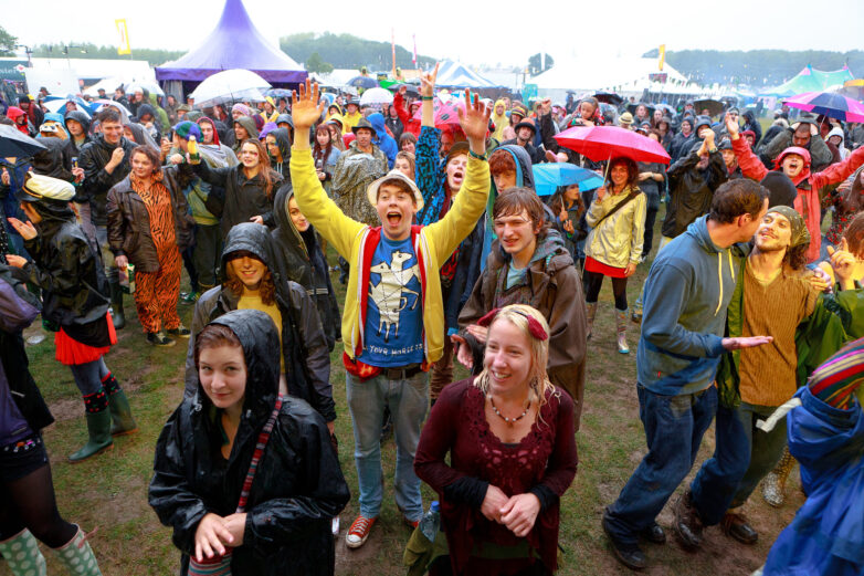A crowd in raincoats and holding umbrellas enjoys an outdoor festival in wet, muddy conditions, reflecting the lively spirit seen throughout Shambala Festival history. Some are cheering, smiling, and clapping, whilst others chat and dance under the overcast sky.