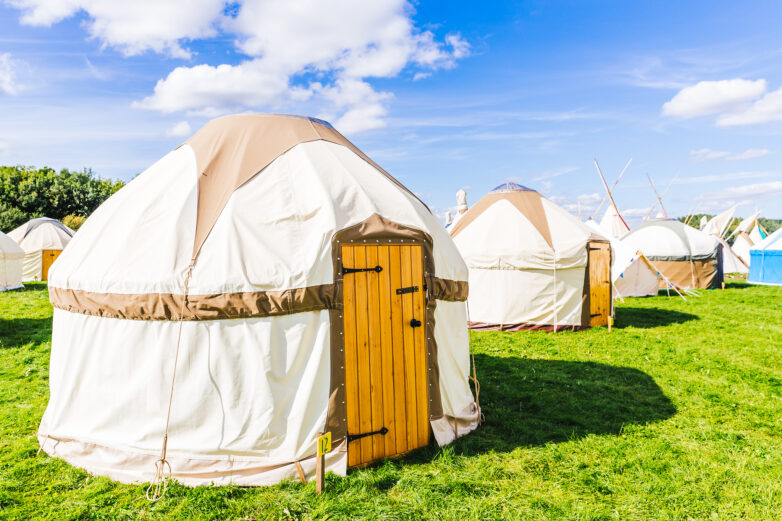 A group of large, round cream and beige tents with wooden doors set up on a grassy field under a bright blue sky with scattered clouds.