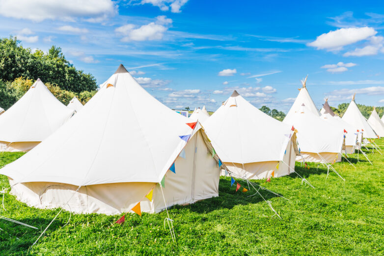Several large white canvas tents are set up on green grass under a bright blue sky with scattered clouds. Colourful pennant flags decorate the tents, suggesting a festive outdoor event or campsite.