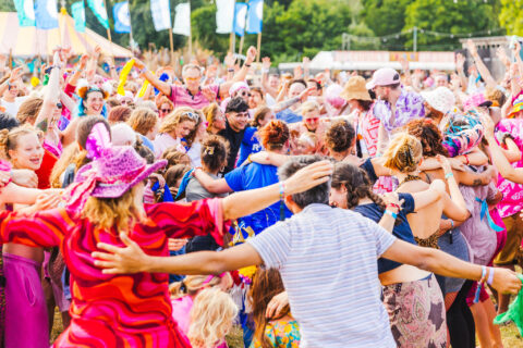 A large, colourful crowd of people at an outdoor festival hug and dance together under bright daylight, with flags and trees in the background.