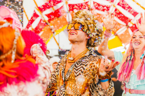 A person wearing a furry hat, animal-print shirt, gold chains, and oversized sunglasses smiles and dances under colourful decorations, surrounded by bubbles and other joyful people in vibrant costumes.