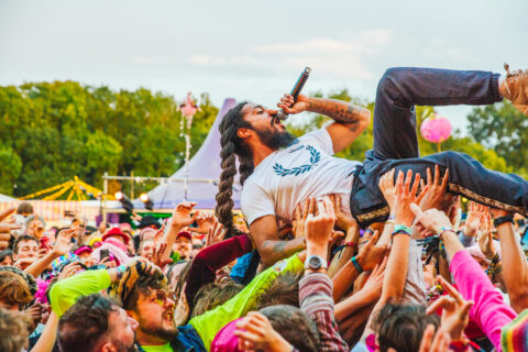 Bob Vylan crowd surfs while performing with a microphone, supported by the hands of excited audience members at Shambala. Trees and festival tents are visible in the background.